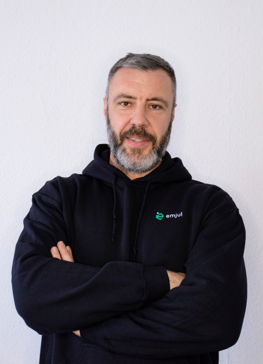 Mann mit grauem Bart und kurz geschnittenem Haar trägt schwarzen Kapuzenpullover mit Emjul-Logo.