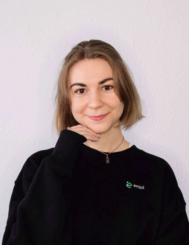 Porträt einer jungen Frau mit kurzem braunem Haar, die schwarz Pullover trägt und lächelt, vor weißer Wand