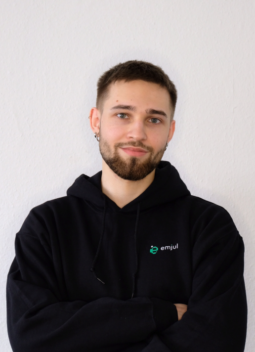 Junger Mann mit kurzem braunem Haar und Bart, trägt schwarzen Hoodie mit Logo vor weißer Wand