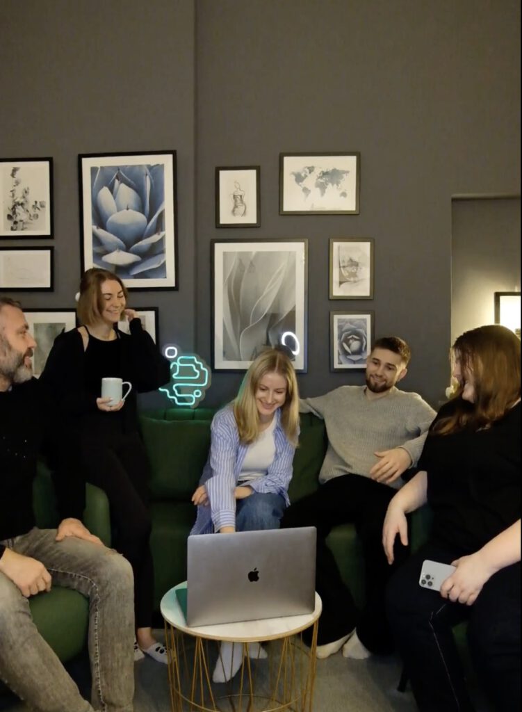 Fünf Personen sitzen und stehen entspannt in einem Wohnzimmer vor einem Laptop auf einem kleinen Tisch.