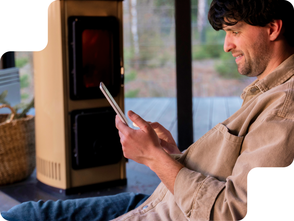 Mann mit rustikalem Hemd sitzt entspannt am Ofen und schaut lächelnd auf sein Smartphone