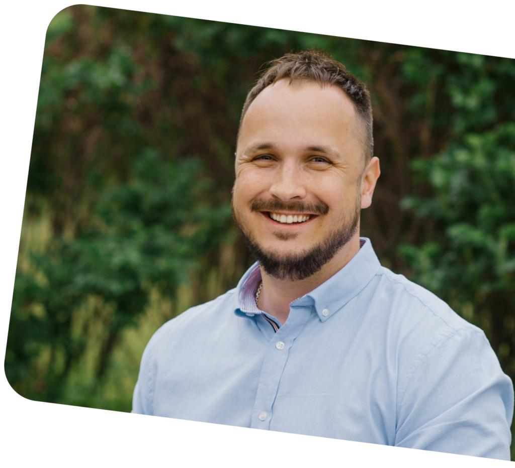 lächelnder Mann mit kurzem braunem Haar und Bart, trägt hellblaues Hemd vor grüner Naturkulisse