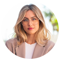 Porträt einer jungen Frau mit blonden Haaren, Brille und beige Jacke vor unscharfem Hintergrund