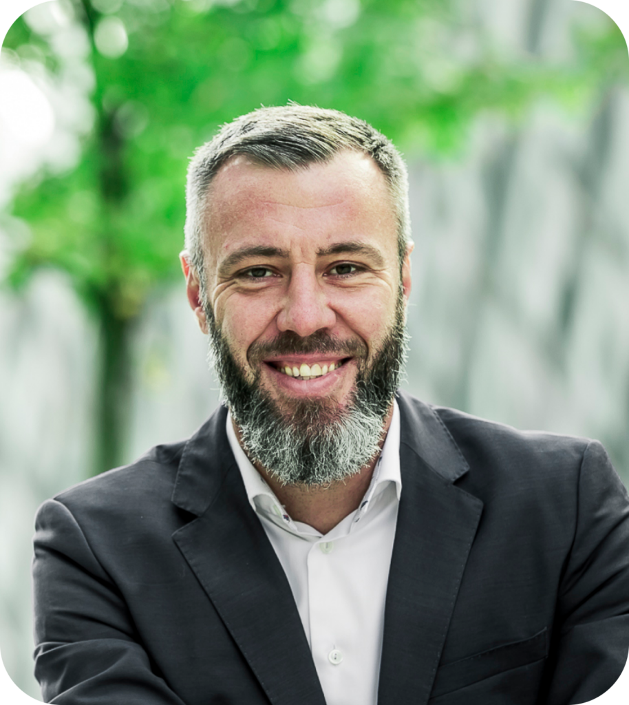 Porträt eines lächelnden Mannes mit grauem Bart im Anzug vor grünem Hintergrund