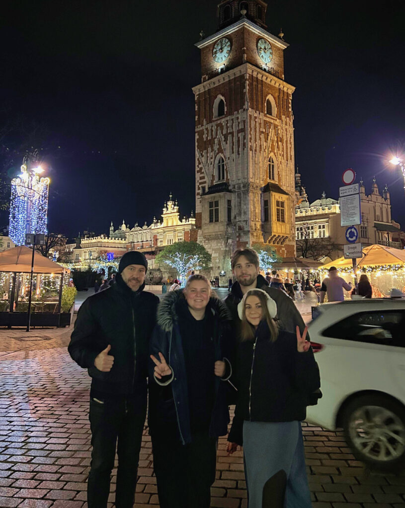Vier Personen posieren vor einem beleuchteten Uhrturm auf einem gepflasterten Platz bei Nacht
