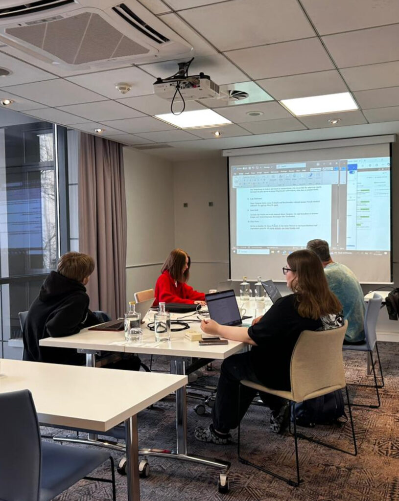 Gruppe von vier Personen in einem Besprechungsraum, beschäftigen sich mit Laptops und Projektion an der Wand.