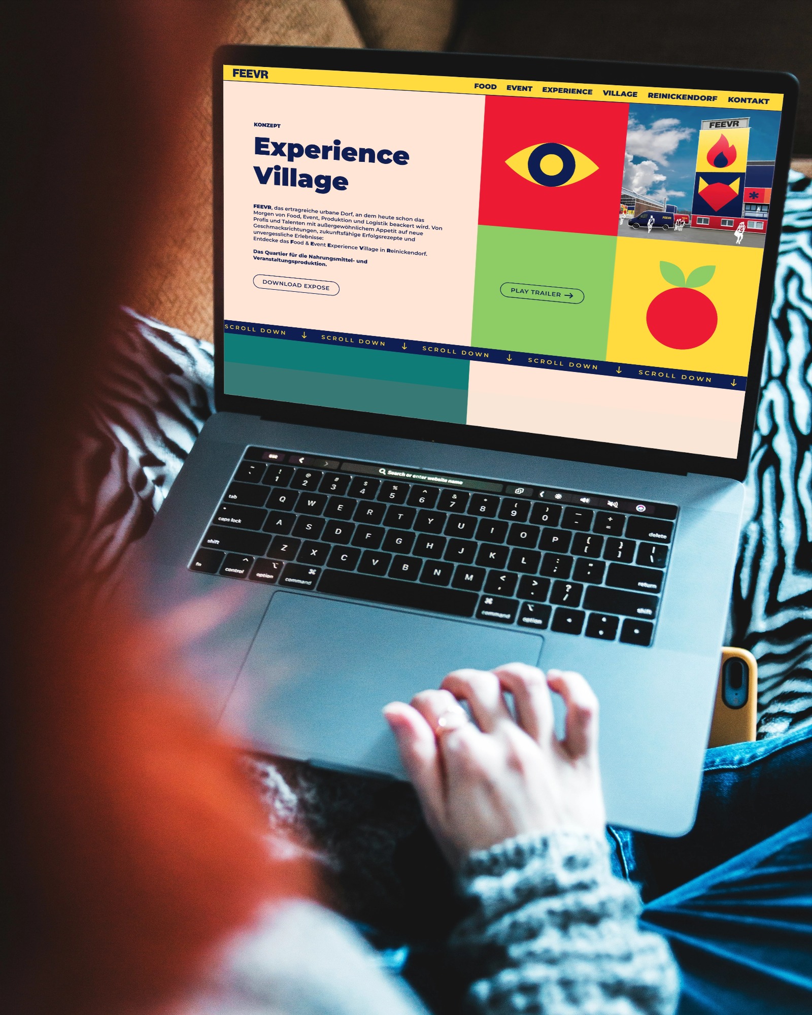 Laptop mit geöffneter Webseite zur Experience Village von FEEVR, mit Abschnitt „Konzept“ und bunten grafischen Elementen, auf dem Schoß einer Person mit rotem Haar und grauem Pullover sitzend.