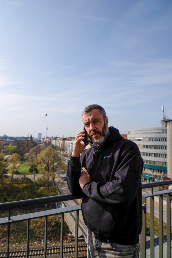 Mann mit Bart in schwarzem Hoodie telefoniert auf Balkon mit Stadtansicht im Hintergrund.