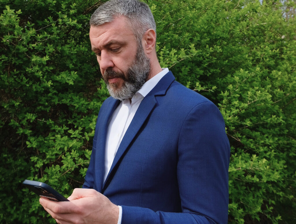 Sergej, der Firmeninhaber, im blauen Anzug, blickt auf sein Smartphone vor einem Hintergrund aus grünem Gebüsch.